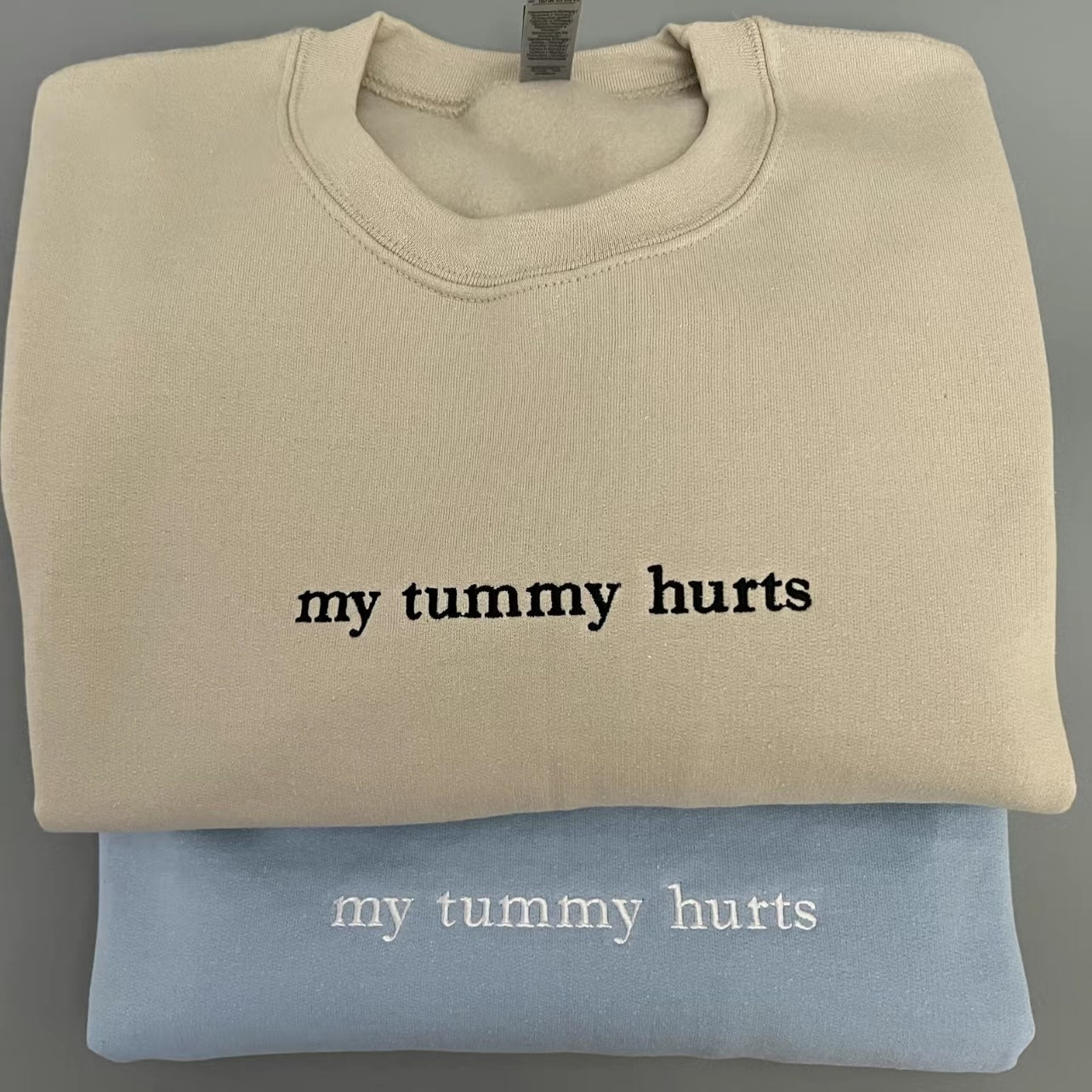 Beige and blue "my tummy hurts" sweatshirts stacked on top of each other.