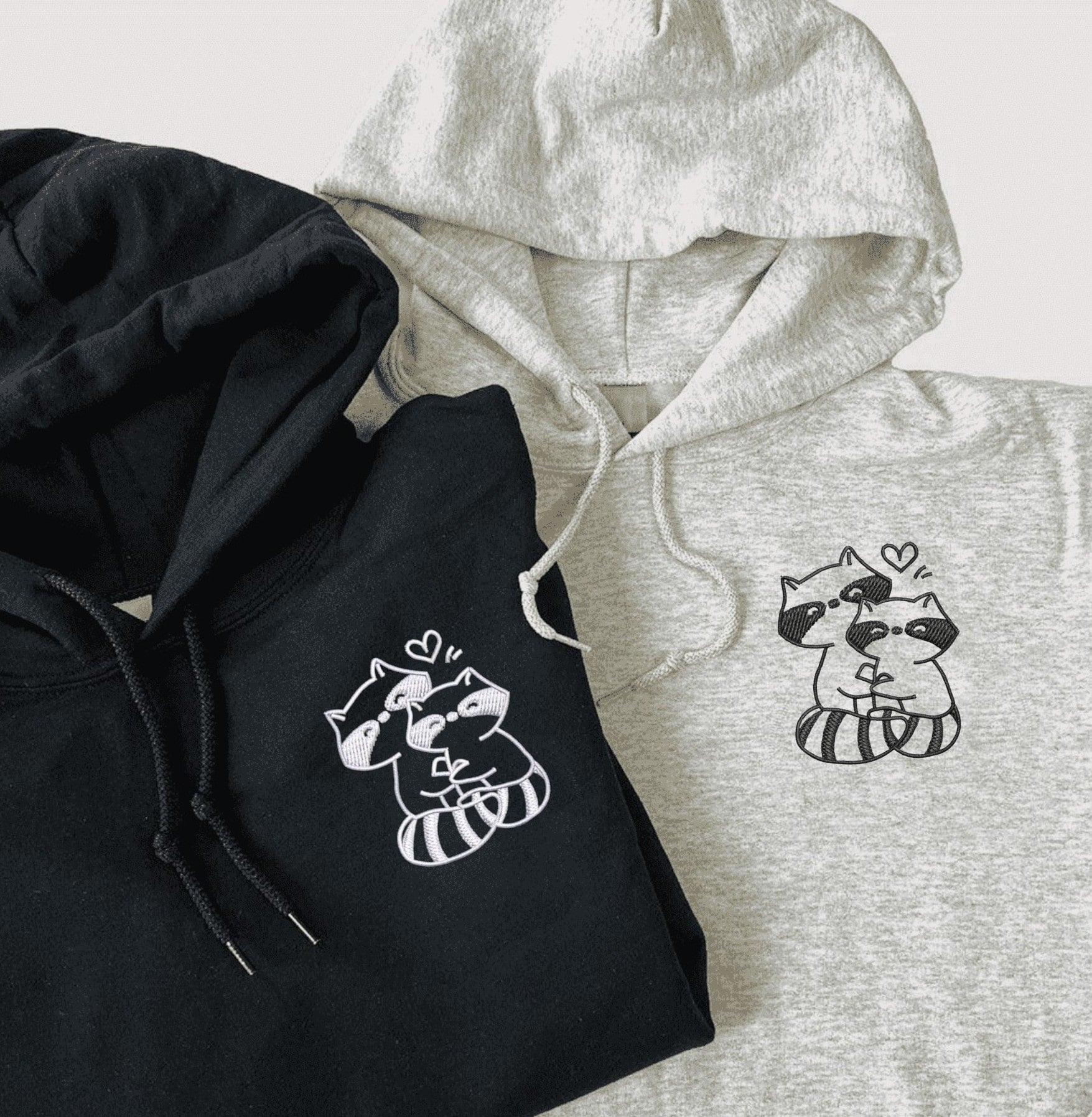 Trash Pandas Couple Hoodies featuring embroidered raccoons hugging with heart designs.