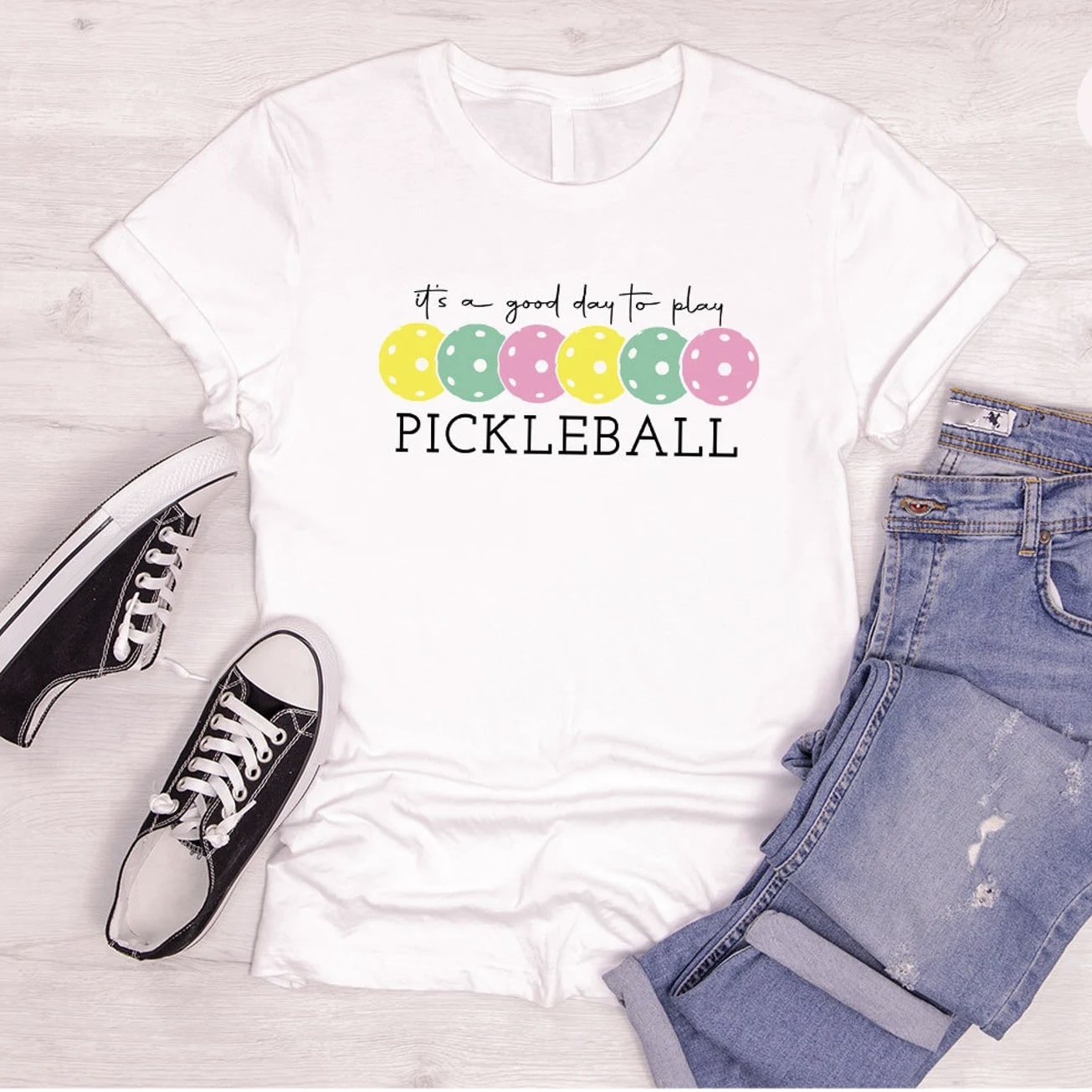 White t - shirt with pickleball design, paired with jeans and sneakers on a wooden floor