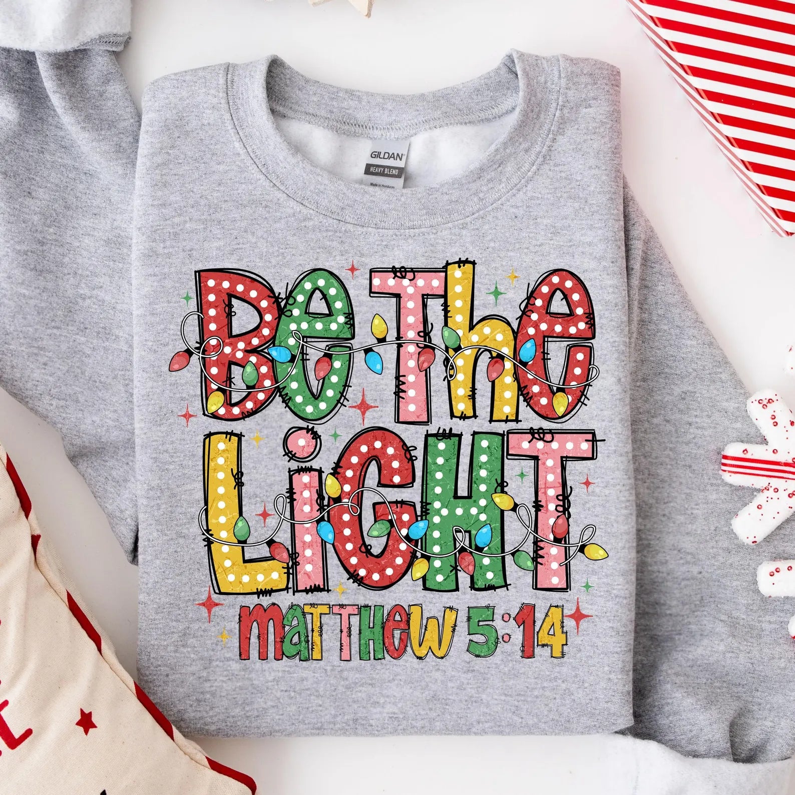 Christian holiday sweatshirt with 'Be The Light' message and bright Christmas lights.