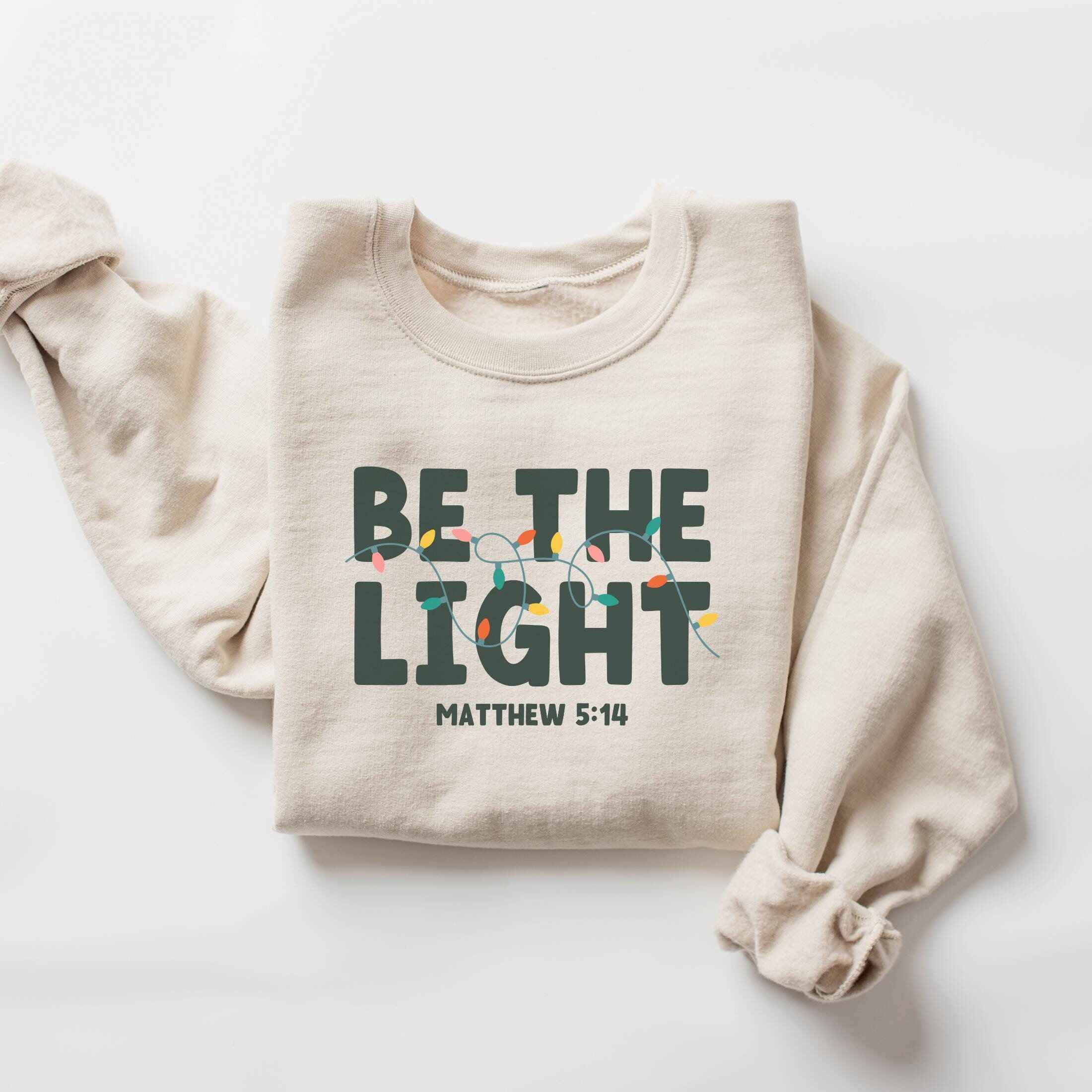 Inspirational Christmas sweatshirt with "Be the Light" design, customizable colors
